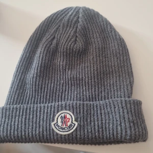 Grå ribbstickad mössa från Moncler - Snygg grå ribbstickad mössa från Moncler med klassisk uppvikt kant och broderad logotyp framtill. Perfekt accessoar för kyliga dagar, tillverkad i mjukt och värmande material. Enkel att matcha med olika outfits och ger en clean look.