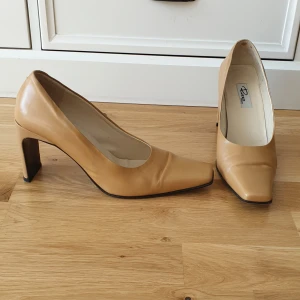 Beige pumps med klack från Rizzo - Stilrena pumps i beige skinn från Rizzo, tillverkade i Italien. Skorna har fyrkantig tå, hög klack och en klassisk siluett som ger en elegant vibe. Perfekta för dig som gillar minimalistisk och tidlös design.