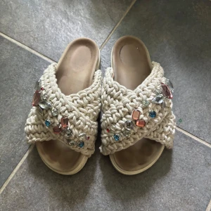 Beige virkade sandaler med stenar - Unika sandaler i beige virkat material med breda korsade band och stora färgglada stenar i blått, rosa och silver. 