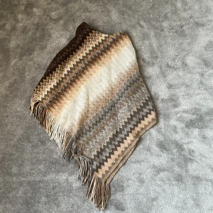 Mönstrad poncho  i bruna toner - Snygg poncho med zigzag-mönster i bruna, beige och grå nyanser. Halsduken har fransar i ändarna och är perfekt för att ge din outfit lite extra stil. Mjuk och skön att ha runt halsen under kyliga dagar.
