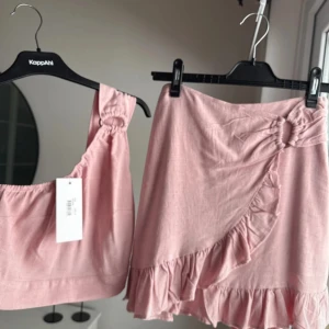 Rosa linnetopp oneshoulder och kjol med inbyggd byxa - Supersöt ljusrosa linnetopp med oneshoulder och snygg ringdetalj på axeln. Toppen är croppad och har en tight passform, perfekt till sommaren. Materialet är linneblandning som känns svalt och fräscht.