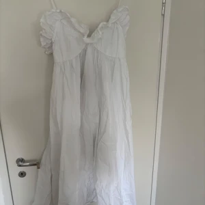 Vit långklänning med volangdetaljer - Superfin vit långklänning med tunna axelband och volang vid bysten. Klänningen har en luftig och flowy passform, perfekt för varma dagar. Tillverkad i bomull som känns lätt och skön mot huden. Söt och romantisk stil med feminina detaljer.