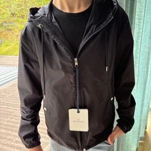 Svart vindjacka från Moncler - Snygg svart vindjacka från Moncler med huva och dragsko. Jackan har dragkedja framtill, två fickor med tryckknappar och elastiska muddar vid ärmslut. Klassisk Moncler-logga på ärmen och stilren design som passar till allt.