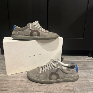 Grå mockasneakers från Oliver Cabell - Snygga grå sneakers i mocka från Oliver Cabell med rund tå och platt sula. Skorna har en diskret cirkeldetalj på sidan och blå detalj vid hälen. Klassisk snörning och stilren design som passar till det mesta.