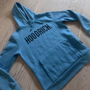 Blå hoodie från Hoodrich - Snygg ljusblå hoodie från Hoodrich med stor svart logga på bröstet. Tröjan har huva med dragsko och en stor magficka framtill. Perfekt för en chill och avslappnad stil. 