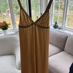 Guldfärgad klänning LiseLotte Westerlund - Elegant klänning i guldfärgad satin från LiseLotte Westerlund. Klänningen har smala axelband, bar rygg och kontrasterande bruna detaljer vid bysten. Materialet är glansigt och faller snyggt längs kroppen.
