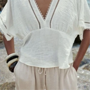Vit blus med spets och v-ringning - Luftig vit blus i bomull med djup v-ringning och fina spetsdetaljer längs halsringningen och ärmsluten. Blusen har korta, vida ärmar och avslappnad passform, perfekt för varma dagar.