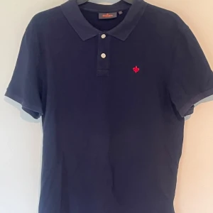 Mörkblå pikétröja från Morris - Snygg mörkblå pikétröja från Morris med klassisk krage och två knappar framtill. Tröjan har korta ärmar och en röd broderad logga på bröstet. Tillverkad i bomull för en skön och luftig känsla. Perfekt för en stilren och avslappnad look.