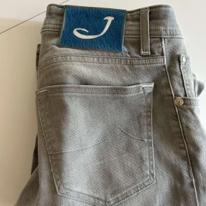 Grå jeansbyxor från Jacob Cohën - Snygga grå jeansbyxor från Jacob Cohën med klassisk femficksdesign och ett blått patch med J-logga bak. Jeansen har raka ben och är tillverkade i mjukt bomullstyg för en skön känsla. Kom privat för fler bilder