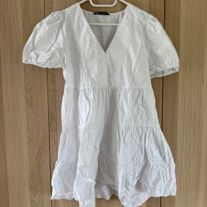 Vit kortklänning med puffärm från Zara - Supersöt vit kortklänning från Zara med v-ringning och puffiga korta ärmar. Klänningen har volangdetaljer och är sydd i ett lätt bomullstyg, perfekt för varma dagar. Enkel och fräsch look som passar till sneakers eller sandaler.