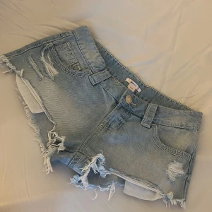 Lågmidjade jeansshorts med slitningar från Gina Tricot - Snygga ljusblå jeansshorts från Gina Tricot med slitna detaljer och fransig kant. Shortsen har klassisk femficksmodell, nitar på bakfickorna och låg midja. Perfekta för sommaren och ger en cool, avslappnad vibe.