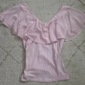 Ljusrosa volangblus offshoulder - Supersöt ljusrosa blus med volang och offshoulder-modell. Toppen har en flowy, luftig känsla och är perfekt för dig som gillar romantiska vibes. Tunt, lätt material som faller snyggt över axlarna. Perfekt till jeans eller kjol för en söt look.