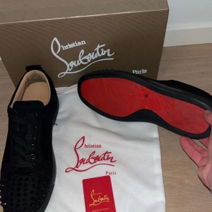 Christian Louboutin svarta sneakers med nitar - Svarta sneakers från Christian Louboutin i mocka med ikoniska röda sulor. Skorna har rund tå, snörning och coola svarta nitar på tån för en edgy vibe. Insidan är beige och sulan är platt. Perfekta för dig som vill sticka ut med lyxig streetstyle.
