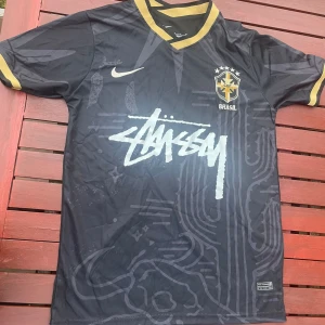 Svart Brasil x Stüssy Nike fotbollströja - Unik svart Brasilien fotbollströja från Nike i samarbete med Stüssy. Tröjan har guldiga detaljer på krage och ärmslut, Brasil-logga på bröstet och Stüssy-tryck framtill. Tillverkad i Dri-FIT-material som andas, perfekt för träning eller streetwear.