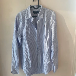 Blårandig skjorta från Polo Ralph Lauren - Klassisk blårandig skjorta från Polo Ralph Lauren i bomull. Skjortan har lång ärm, knappar framtill och broderad logga på bröstet. Snygg krage och klassisk passform som funkar till många olika stilar.