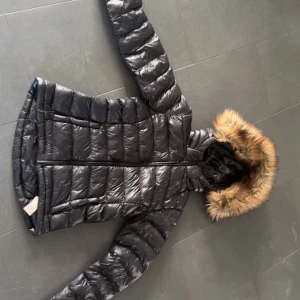 Svart dunjacka med fuskpäls från Everest - Snygg svart dunjacka från Everest med glansig finish och huva med fuskpäls. Jackan har långa ärmar, dragkedja framtill och quiltad design som ger en cool look. Perfekt för kalla dagar när du vill hålla stilen.aldrig andvänd! I storlek 158-164