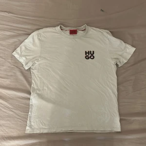 Vit t-shirt från Hugo Boss - Vit t-shirt från Hugo Boss med svart och röd HUGO-logga på bröstet. Klassisk rund hals och korta ärmar. Skön och enkel stil som funkar till allt. Tillverkad i mjuk bomull för en bekväm känsla.