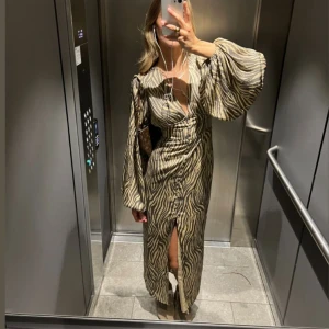 Zebramönstrad långklänning med v-ringning - Snygg långklänning med zebramönster i beige och grå toner. Djup v-ringning, långa puffärmar och knappar framtill hela vägen ner. Klänningen har en slits framtill och markerad midja för en trendig siluett.