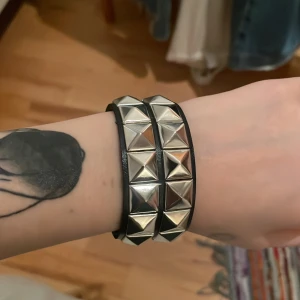 Svart nitar armband med silverdetaljer - Coolt svart armband med dubbla rader av stora silvriga pyramidnitar. Perfekt accessoar för en edgy stil och passar lika bra till jeansjacka som skinnjacka. Armbandet har en bred design och ger en tuff rockig känsla.