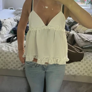 Vit volangtop med smala axelband - Supersöt vit topp med smala axelband och v-ringning. Toppen har volangdetaljer både under bysten och längst ner, vilket ger en luftig och romantisk känsla. Perfekt till jeans eller kjol för en fräsch sommarlook.