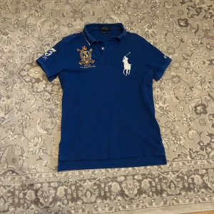 Blå pikétröja Polo Ralph Lauren S - Snygg blå pikétröja från Polo Ralph Lauren med broderad logga och stort vitt polospelare-märke på bröstet. Klassisk krage med vita detaljer och knappar framtill. Broderad gul detalj och nummer 3 på ärmen. Perfekt för en sportig och stilren look.
