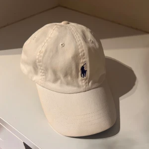 Keps Polo Ralph Lauren - Tidlös vit keps från Polo Ralph Lauren med  Justerbart spänne i brunt läder för perfekt passform. Kepsen är i bomull och har en stilren design. Kepsen är ren och fräsch då den blivit tvättad innan försäljning 🫧🧼✨