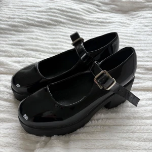 Svarta Mary Jane ballerinaskor med klack, helt oanvända!! - Snygga svarta Mary Jane ballerinaskor från Koi med lackad finish och bred rem med silverfärgat spänne. Skorna har rund tå, plattformsula och låg klack som ger extra attityd. Perfekta för dig som vill ha en edgy men söt look.