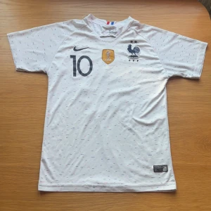 Frankrike Mbappé 10 VM 2018 Nike tröja - Säljer en officiell Frankrike fotbollströja från VM 2018 med Mbappé och nummer 10 på ryggen. Tröjan är vit med små blå och röda streck, har korta ärmar och är tillverkad i Dri-Fit material för bästa komfort. Nike-logga och franska landslagets emblem på bröstet.