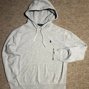 Ralph Lauren Hoodie - Snygg ljusgrå hoodie från Polo Ralph Lauren med klassisk broderad logga på bröstet. Den är i bra skick men lite sliten vid ändarmen som man ser på sista bilden.