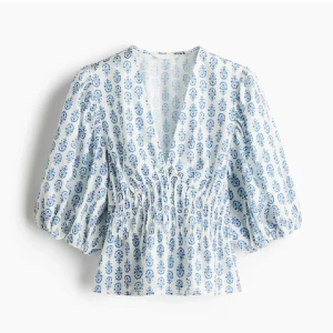 Hm blus - Superfin blus i vitt med blått blommigt mönster och v-ringning. Modellen har puffiga trekvartsärmar och smockad midja som ger en snygg siluett. Perfekt till jeans eller kjol för en fräsch look. Lätt och luftig känsla, perfekt för varmare dagar.