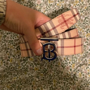 Rutig Burberry bälte med B-spänne - Snyggt bälte från Burberry med klassiskt rutigt mönster i beige, brunt och rött. Bältet har ett stort metallspänne med Burberrys ikoniska B-logga i silver. Perfekt accessoar för att lyfta din outfit med en lyxig touch.