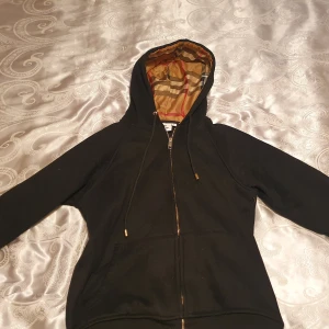 Svart Burberry hoodie med rutig huva - Svart hoodiejacka från Burberry med dragkedja och klassisk rutig huva i beige, rött och svart. Jackan har snörning i huvan, känguruficka framtill och är helsvart i kroppen. Perfekt för dig som vill ha en stilren men ändå lyxig look.