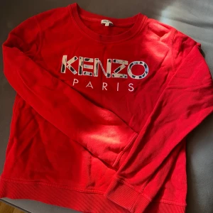 Röd Kenzo sweatshirt med broderad logga - Röd sweatshirt från Kenzo med broderad logga i vitt, beige, blått och svart framtill. Tröjan har rund halsringning, ribbade muddar och är långärmad. Perfekt statement-piece för dig som gillar streetstyle och vill sticka ut.