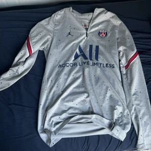 PSG långärmad fotbollströja grå - Snygg långärmad PSG-tröja i ljusgrått med mörkblå stänk, blå logga och tryck. Röd och blå rand på ärmarna, dragkedja vid halsen och klassisk PSG-emblem på bröstet. Tillverkad i lätt och ventilerande material, perfekt för träning eller match.