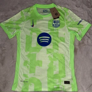 Barcelona x Nike fotbollströja  - Säljer en FC Barcelona fotbollströja från Nike i neonljusgrön färg med abstrakt mönster. Tröjan har korta ärmar, klubbmärke på bröstet, blått Spotify-tryck och UNHCR-logga på ryggen. Tillverkad i lätt och ventilerande Dri-Fit material, perfekt för match eller träning. Även perfekt för en stilren look