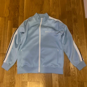 Ljusblå track jacket från Palm Angels(tagen finns oxå) - Säljer en ljusblå track jacket från Palm Angels med vita och svarta ränder längs ärmarna. Jackan har dragkedja framtill, ståkrage och två fickor med dragkedja. Säljer den eftersom jag har haft på mig den en gång men den är liten på mig så måste få bort den.