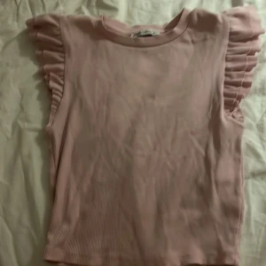 Rosa  sommar volangtopp från Zara - Supersöt ljusrosa topp från Zara med ribbad struktur och volangdetaljer på axlarna. Ärmlös modell som ger en trendig och feminin look. Perfekt att styla med jeans eller kjol för en fräsch vibe.