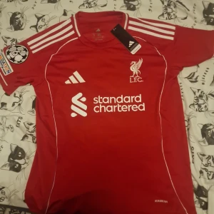 Liverpoo 25/26l röd matchtröja Adidas Wirtz 17 - Liverpool FC matchtröja i rött från Adidas med vita detaljer och tryck. Kortärmad med Champions League-märke på ärmen, klubbmärke och sponsortryck fram samt Wirtz 17 på ryggen. Tillverkad i lätt funktionsmaterial (Aeroready) för bästa komfort. Är helt oanvänd och tvätta den rör hand varsamt så att inte trycket försvinner eller förstörs 