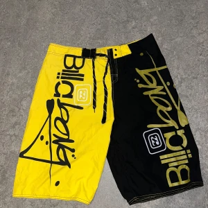 Billabong badshorts i svart och gult - Feta badshorts från Billabong i tvådelad design, ena benet är knallgult och andra svart. Stora loggor på båda sidor och snörning i midjan. Perfekta för stranden eller poolen, med bakficka och cool streetstil.
