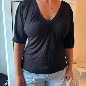 Svart v-ringad topp från H&M - Svart topp från H&M i storlek L med v-ringning och lätt rynkade detaljer vid halsen. Modellen har halvlånga ärmar och är tillverkad i mjuk bomull, perfekt för en chill och enkel look.