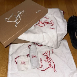 Christian Louboutin svarta sneakers med nitar - Svarta sneakers i mocka från Christian Louboutin med ikonisk röd sula och svarta nitar på tån. Skorna har snörning och rund tå, samt lyxig känsla med läderfoder. Perfekta för dig som vill sticka ut med edgy detaljer och exklusiv design.