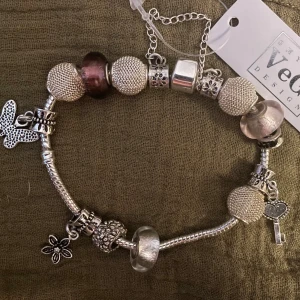 *NY*Silverfärgat charmarmband med berlocker fr VeGi Design  - Charmigt armband i silverfärg från VeGi Design med flera detaljerade berlocker, bland annat fjäril, blomma och hjärtformad nyckel. Armbandet har glittriga och släta pärlor samt inslag av transparenta och ljust rosa/lila pärlor. Skickas i smyckespåse.  För stort för mig som har väldigt smala handleder, passar bäst på ngn med ”normalstora” handleder.