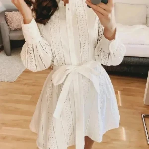 Vit kort klänning med knytband - Superfin vit kortklänning med broderade detaljer och knytband i midjan. Klänningen har långa puffärmar och knappar framtill. Perfekt för sommarens alla tillfällen och ger en fräsch och romantisk look.