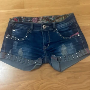 Mörkblå jeansshorts med nitar - Snygga mörkblå jeansshorts med slitna detaljer, uppvikta ben och coola nitar längs kanten. Fickor fram och bak, samt färgglatt mönstrat innerfoder i midjan. Vet inte vilket märke det är. Va originellt köpta för 1200kr. Priset kan förhandlas!!