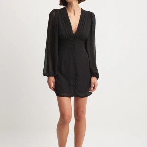 Svart kort klänning med knappar - Supersnygg svart kort klänning med v-ringning och långa, transparenta ärmar. Klänningen har markerad midja och dekorativa knappar framtill hela vägen ner. Perfekt för dig som gillar en stilren och elegant look. OANVÄND MED PRISLAPP 