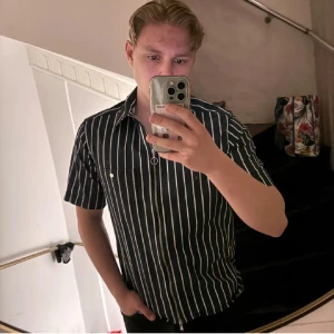 Svartvit randig skjorta J.Lindeberg - Svart skjorta från J.Lindeberg med vita vertikala ränder, kort ärm och dragkedja framtill. Skjortan har klassisk krage och en bröstficka med knapp. Perfekt för dig som gillar en stilren och modern look.