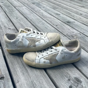 Golden Goose vita sneakers med stjärna - Snygga vita sneakers från Golden Goose med beige mockadetaljer och den ikoniska stjärnan på sidan. Skorna har snörning, rund tå och en vintageinspirerad sula. Perfekta för dig som gillar en avslappnad och trendig stil.