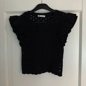 Svart stickad topp med volangärmar från Zara - Svart stickad topp från Zara med korta volangärmar och hålmönster. Toppen har en croppad passform, längd fram 32cm. Två trådar som öglat sig, se bilder. 