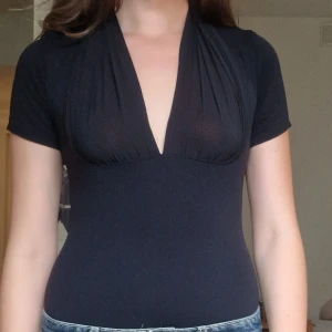Svart v-ringad body med kort ärm - Säljer en svart, tight body med djup v-ringning och korta ärmar. Toppen har en smickrande passform och lätt rynkade detaljer vid bysten. 