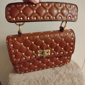 Brun quiltad handväska med nitar - Säljer Valentino liknande brun quiltad handväska med guldiga nitar och snyggt spänne framtill. Väskan har ett brett handtag och kan användas som både Axel och handväska. 